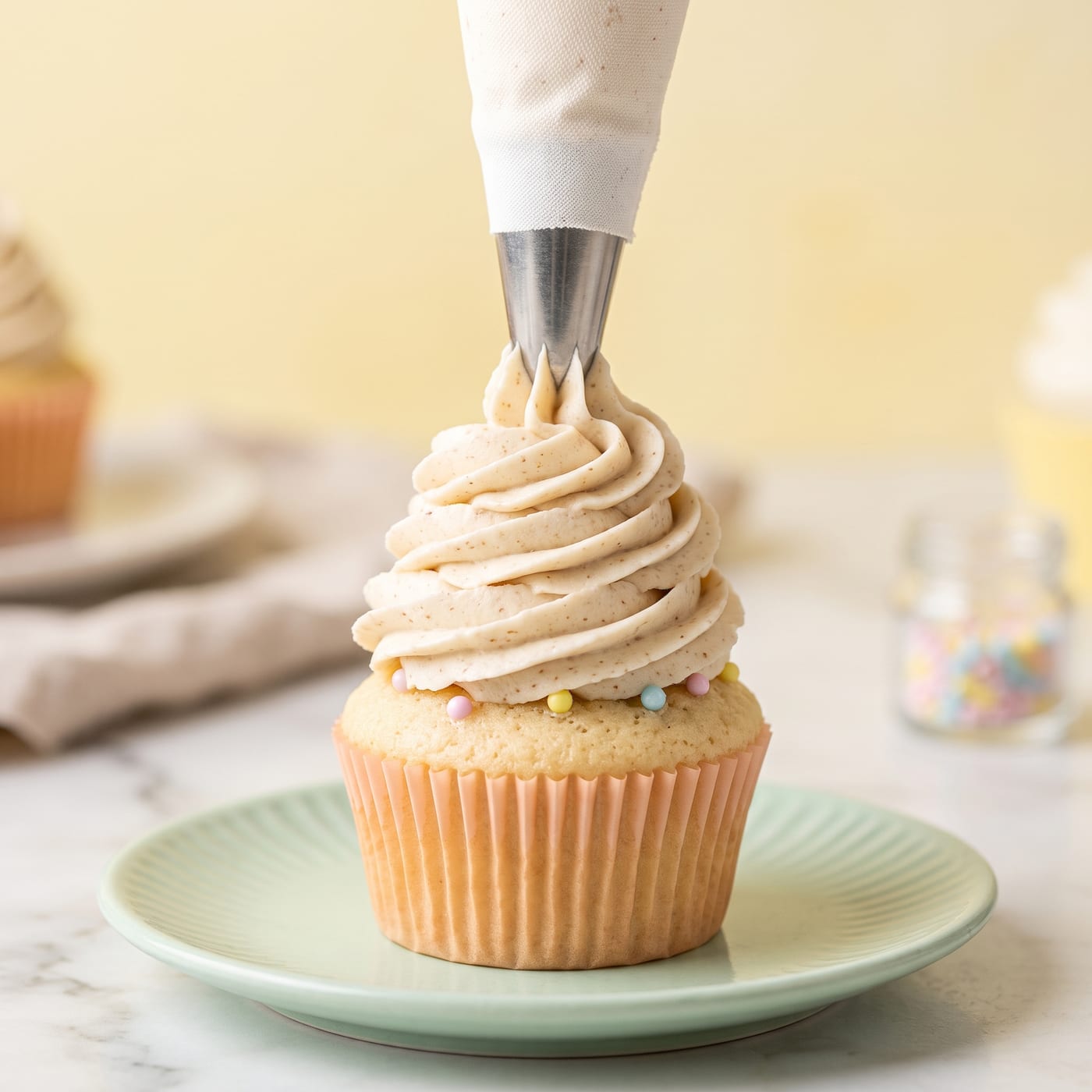 Almond buttercream being piped onto a cooled vanilla cupcake with a star tip