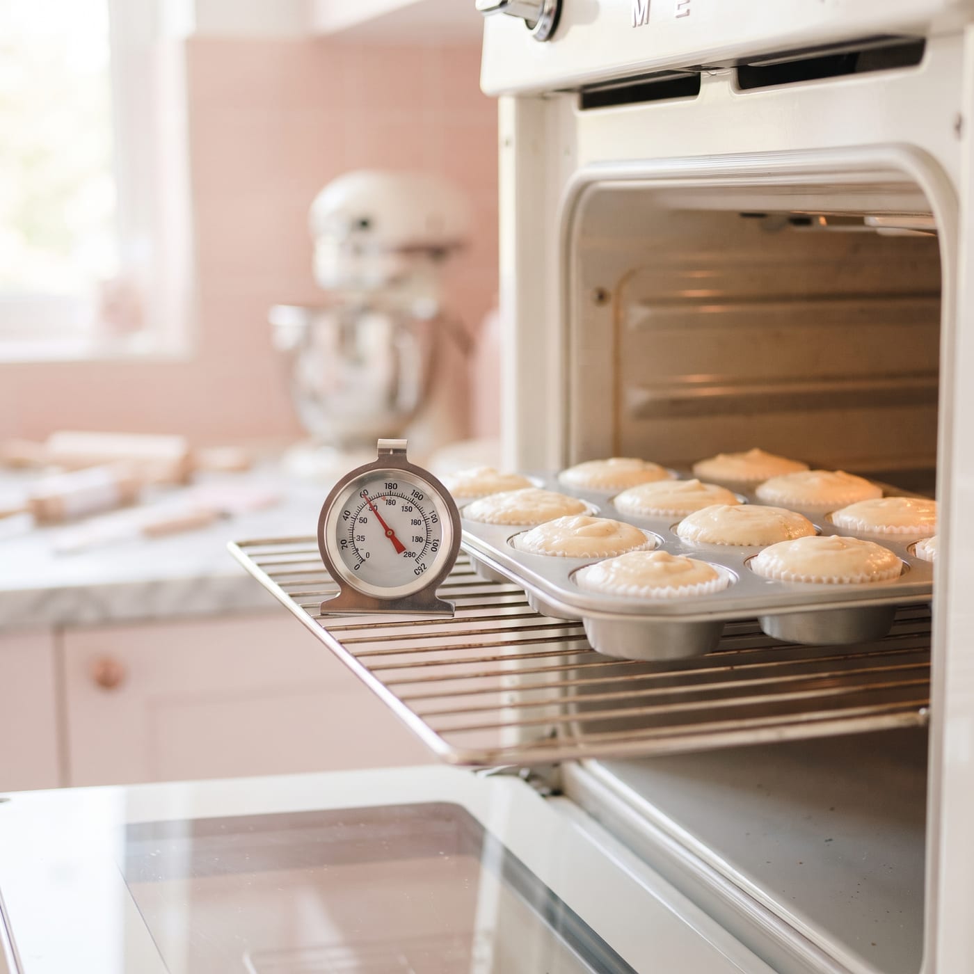 Close up of an oven thermometer resting inside an open oven next to a cupcake pan filled with batter