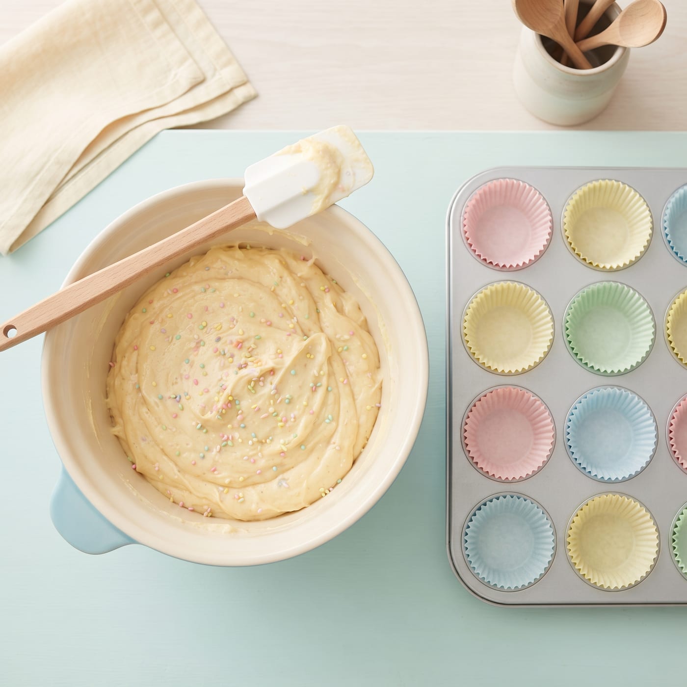 Bowl of light yellow cupcake batter with naturally colored sprinkles folded in, next to a lined muffin pan
