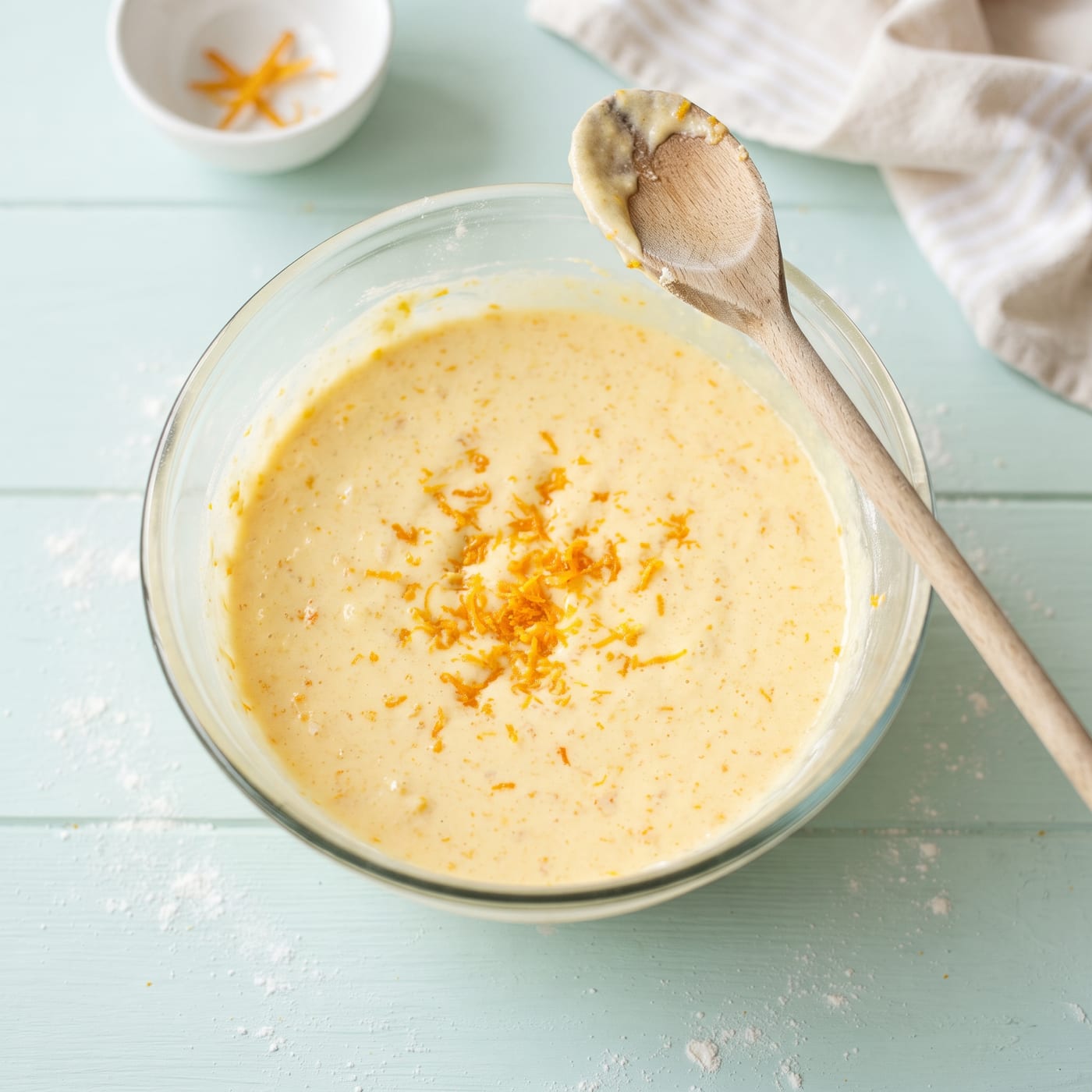 Orange zest being folded into pale cupcake batter in a glass mixing bowl
